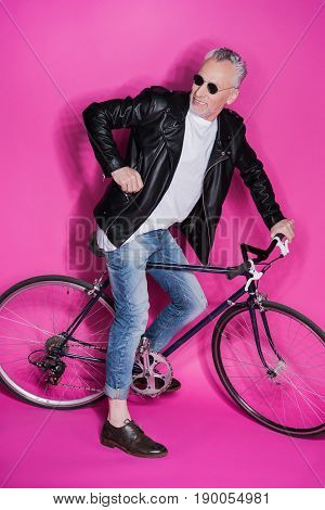 Stylish Senior Man Wearing Leather Jacket, Sunglesses And Riding Bicycle Isolated On Pink
