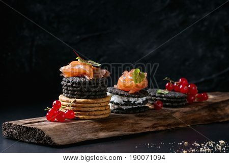 Stack of black charcoal and traditional crackers with smoked salmon, cream cheese, green salad and red currant berries on ood serving board over black metal background. Appetizer snack. Space for text