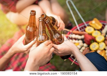 Group of friends clinking bottle of beer during camping outdoor party with barbecue in background