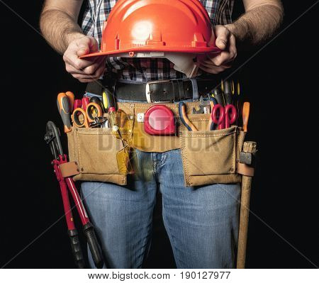 detail of handyman with leather toolsbelt and tools on dark background holding helmet