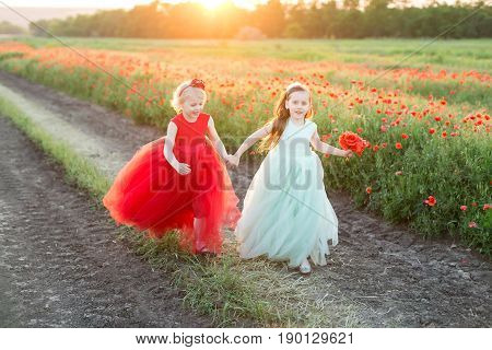 freedom, frendship, country, happiness concept - in gold light of sun two lovely girls in beautiful feast dresses in bright red and light blue shadows running in rural road near the field of poppies