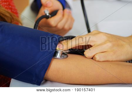 Close up of a young doctor using the stethoscope to hear the pulse while a woman is with a tensiometer in her arm, in a doctor consulting room background.