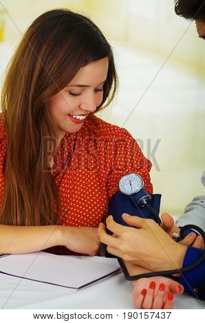Beautiful young patient using a manual tensiometer in her arm, in a doctor consulting room background.