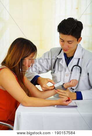 Close up of a handsome smiling young doctor with stethoscope around his neck taking the pulse to a young woman with a tensiometer, in a doctor consulting room background.