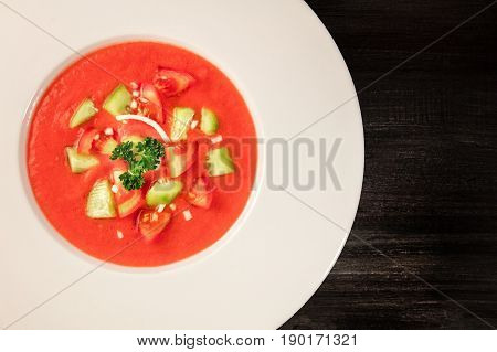 A closeup of a plate of gazpacho soup with slices of tomato, cucumber, and onion, with parsley leaves, on a dark background with a place for text