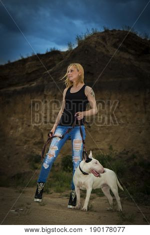 Brutal, bold girl with a fighting dog breed of bull terrier on a background of a rock in the evening