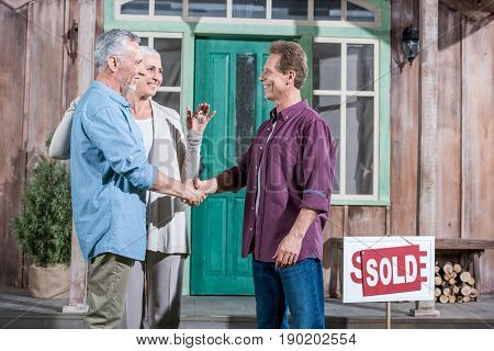 Senior Wife Holding Keys Of New House While Her Husband Shaking Hands With Saleman