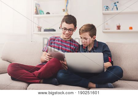 Technology, boy friendship and communication concept - two cute teenagers using tablet and laptop while sitting on the couch in living room at home