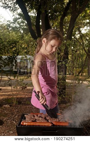 Little caucasian girl (elementary age) cooking the ćevapi (a grilled dish of minced meat a type of skinless sausage) on barbecue grill outdoor. Summer camping active recreation and touristic concept.