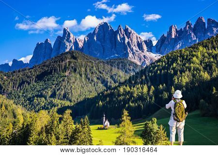 Active elderly woman-tourist with backpack photographs the church of Santa Maddalena in Dolomites. Forested mountains surrounded by green Alpine meadows. The concept of ecological and active tourism