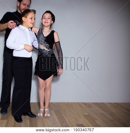 The young boy and girl with male coach or teacher posing at dance studio on gray. The ballroom dancing school and education concept