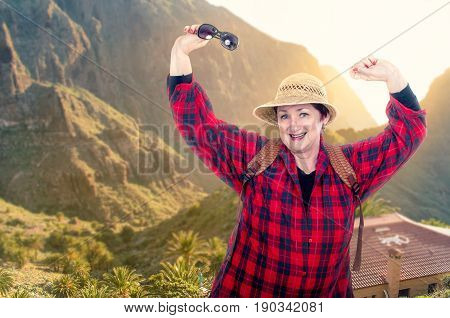 Tireless older woman in yellow straw hat backpacking. She wearing red plaid shirt poses on blurred mountains background are flooded by sunrise. Her right hand holds sunglasses. Older people traveling concept.