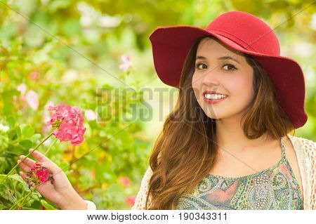 Close up of a cheerful young woman standing outdoors in garden wearing a red hat in garden background.
