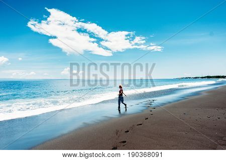 A tall happy red-haired caucasian girl running along a deserted beach along the surf line. Tropical beach with black vulcano sand and blue sky with clouds. Happiness and freedom concept. Bali Indonesia