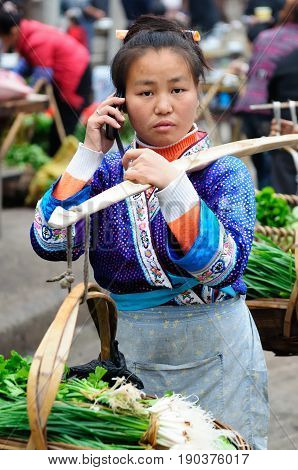 RONGJIANG CHINA - 13 NOVEMBER 2010: Chinese in the ethnic dress carrying the agricultural produce on the bamboo stick which is trying to sell in the city talking through the mobile phone