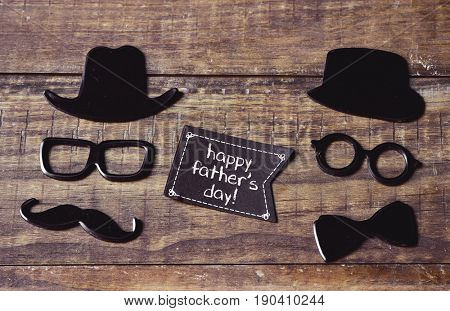a black flag-shaped signboard with the text happy fathers day and two pairs of eyeglasses, a mustache, a bow tie and two hats forming two different men faces, on a rustic wooden surface
