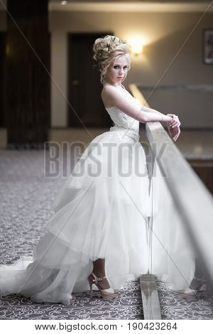 A beautiful bride with a voluminous hairdo with fresh flowers bright makeup in a white lace dress with a train is folded her hands on the railing and looks with a serious look.