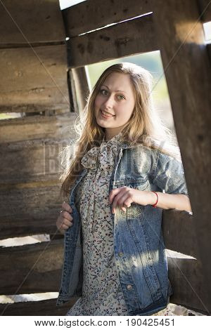 Beautiful girl with green eyes and long blond hair smiling and standing against the old wooden wall and window. The girl is dressed in a white dress with a floral pattern and a light denim jacket.