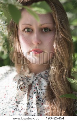The face of a beautiful girl with long slightly wavy blond hair large bright green eyes and long black resils the girl is dressed in a white dress with a bow on her neck with a floral pattern against the background of green leaves of a tree.