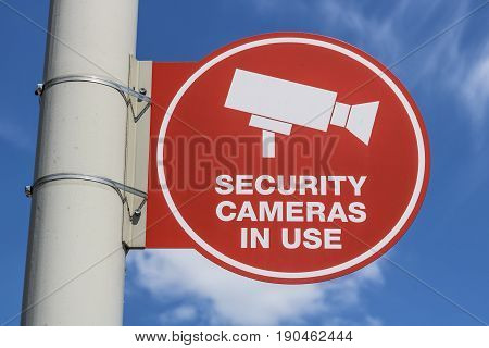 Red Security Camera In Use Sign on Gray Post and Mostly Sunny Background V