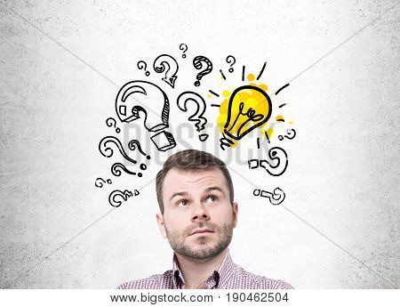 Pensive bearded young man standing near a concrete wall looking upwards. There is a yellow light bulb sketch surrounded by question marks