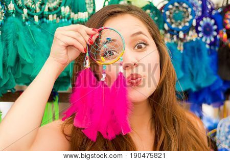 Close up of a beautiful smiling young woman pointing in from of her face a pink catchdreamers, in colorful market fabrics background.