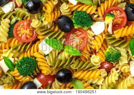 A closeup photo of a pasta salad. Rotini of various colours, fresh green basil leaves, black olives, mozzarella cheese slices, broccoli, and cherry tomatoes