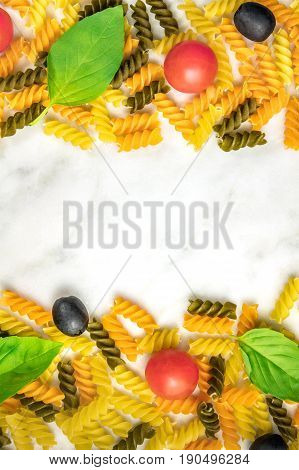Pasta salad ingredients on white marble with a place for text. Rotini, cherry tomatoes, fresh basil leaves, and black olives, forming a frame for copy space