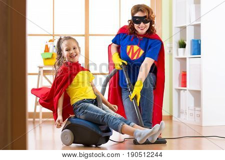 Kid girl and her mother weared Superhero costumes. Cute helper child and woman make cleaning room and have a fun pastime.