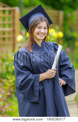 Smiling young woman holding diploma and wearing cap and gown outdoors looking at camera. Graduation concept.