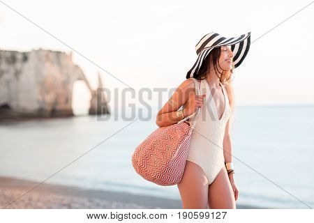 Young woman in hat and swimsuit walking with beach bag near the ocean during the summer vacation