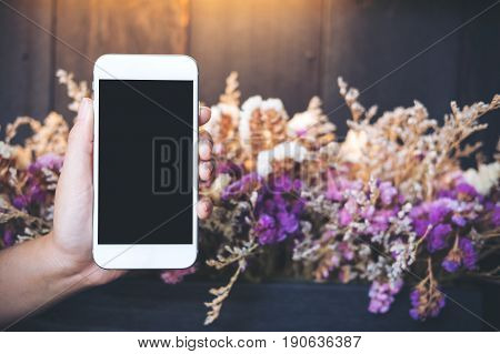 Mockup image of hands holding and showing white mobile phone with blank black screen with colorful dry flowers and wooden wall background in cafe