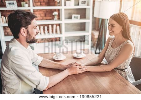 Couple in love is holding hands on wooden table and look on each other. Cups of coffee are on the background.