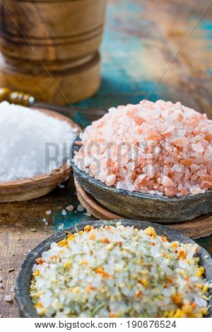 Different types of natural salt in stone bowls on wooden surface. White sea salt pink Himalayan salt Spiced salt with rosemary