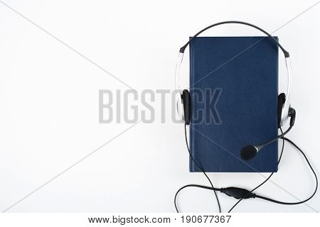 Audiobook on white background. Headphones put over blue hardback book, empty cover, copy space for ad text. Distance education, e-learning concept