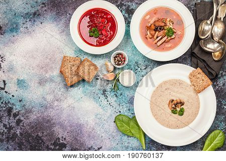 Set of three bowls with different soups: meat soup solyanka, Russian borsh and mushroom cream soup on stone background, top biew.