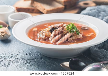 Soup with meat, olives, herbs, lemon in white bowl, black bread and spices on gray background, homemade food. Traditional Russian soup - solyanka
