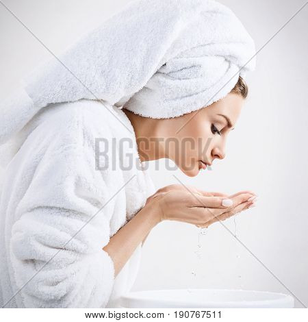 Young woman washing face with clean water in white bathrobe and towel. Morning hygiene concept.