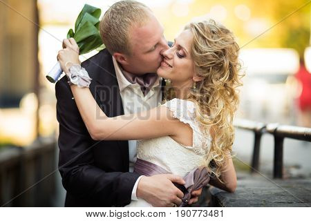 Stylish Groom Kisses A Tiny Blonde Bride Posing On The Bridge