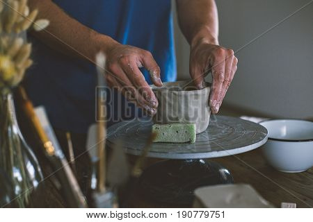 Close up of male hands working with clay on artisan working stand from dry wood surrounded by artistic workshop tools and implements creates poterry diy