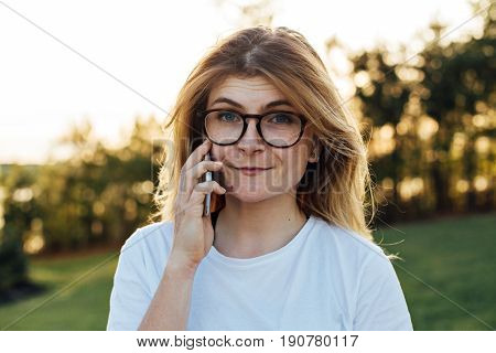 Cute blonde woman with freckles and nerdy goofy funny round glasses chats on phone with her friend or awaits someone to pick up line in soft yellow summer sunset light