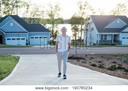 Attractive american teenager in grey sweatpants and white tshirt walks the driveway to a typical residential home on warm summer evening with soft sunset light