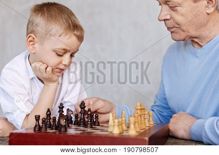 Training problem solving. Clever attentive nice guy teaching his grandson plying chess while he listening him attentively