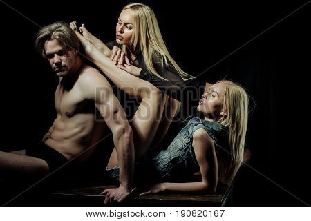 Love triangle. Polygamy and polyamory. Sexy man or macho with muscular body torso and two pretty girls or cute women stylish models with long blond hair on black background. Desire and seduction