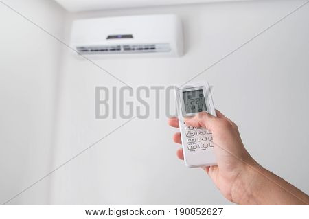 Hand holding remote control, adjusting temperature of air conditioner mounted on a white wall. Indooor comfort temperature