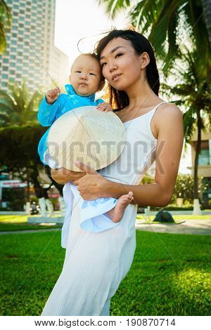 A mother holds her little daughter in traditional vietnam clothers aodai, national costume in her arms on the beach near palms.