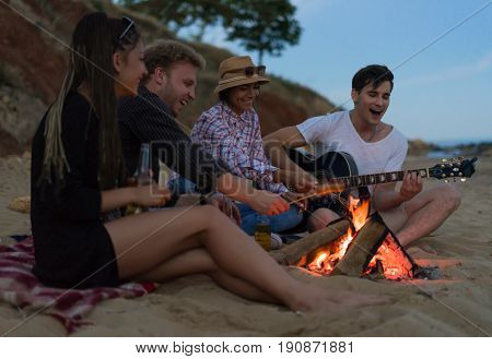 young and cheerful friends sitting on beach and fry sasuages or weenies in bonfire One man is playing guitar. Music on Wild beach