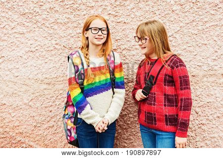 Group of two adorable kid girls posing outdoors against pink wall wearing glasses school backpacks and bright colorful pullovers back to school concept