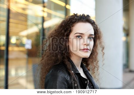 Close-up portrait of mindful Caucasian young attractive woman with dark curly hair staring at camera posing outside. Outdoor shot of confident good-looking girl with a serious thoughtful expression