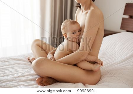 Young attractive nude mom breastfeeding hugging her newborn baby smiling sitting on bed at home. Copy space.